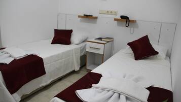 Habitación de hotel con dos camas individuales, cada una con almohada roja y ropa de cama blanca. Hay una mesita de noche pequeña con un libro y un teléfono.