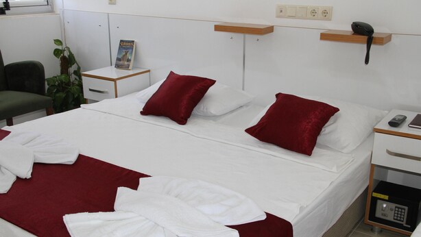Una cama bien hecha con ropa blanca y roja, una mesita de noche con un teléfono y un libro, y una silla verde.