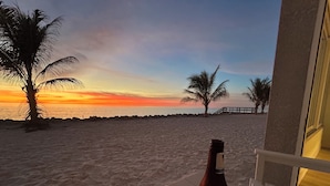 A beach view with a sunset, palm trees, and a bottle on a chair. - Beachfront Ground Level Suite - Everything is Modern & New (Siesta Key)