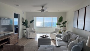 A spacious living room with a large window overlooking the ocean, a ceiling fan, and a flat-screen TV mounted on the wall. - Beachfront Ground Level Suite - Everything is Modern & New (Siesta Key)