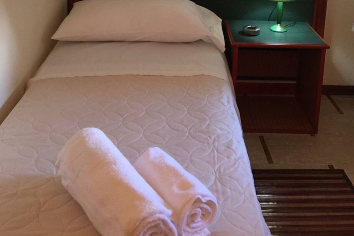 Una cama bien hecha con una almohada, una mesita de noche verde con una lámpara y una toalla enrollada sobre la cama.
