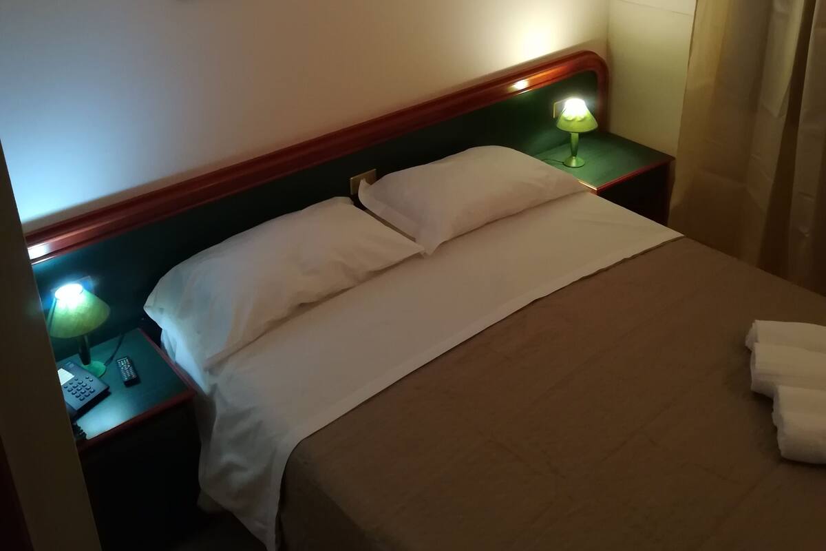 Una habitación de hotel con una cama, dos lámparas de noche, un cuadro en la pared y una ventana con cortinas.