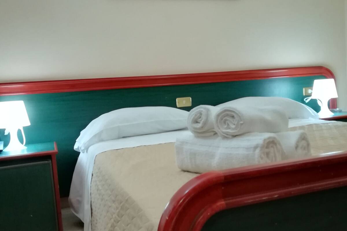 Habitación de hotel con cama, cabecera verde, ropa de cama blanca, un cuadro de una puesta de sol y una mesita de noche con lámpara.
