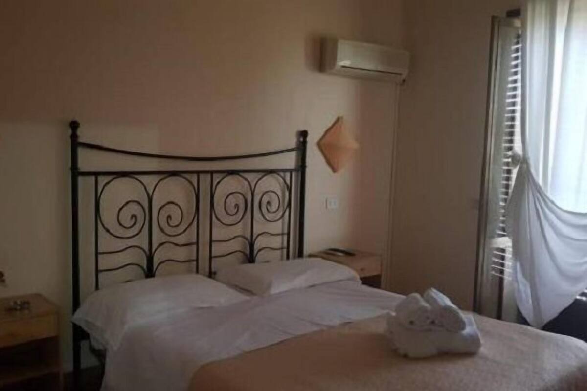 Una cama bien hecha con sábanas blancas, una mesita de noche con lámpara y un aire acondicionado de pared.