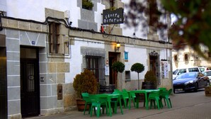 Un edificio con un letrero que dice "Café Ousteria" y una terraza con sillas y mesas de plástico verde.