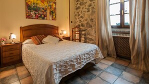 Un dormitorio con una cama grande, una mesita de noche con lámpara, cabecero de madera, pared de piedra y una ventana con cortinas.