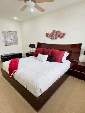 A bedroom with a large bed, a wooden headboard, a nightstand with a lamp, a wall-mounted artwork, and a ceiling fan.