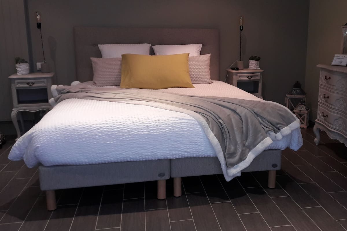 A neatly made bed with a white duvet, a gray throw blanket, and a yellow pillow. There are two bedside tables with lamps, a dresser, and a small nightstand with a decorative item.