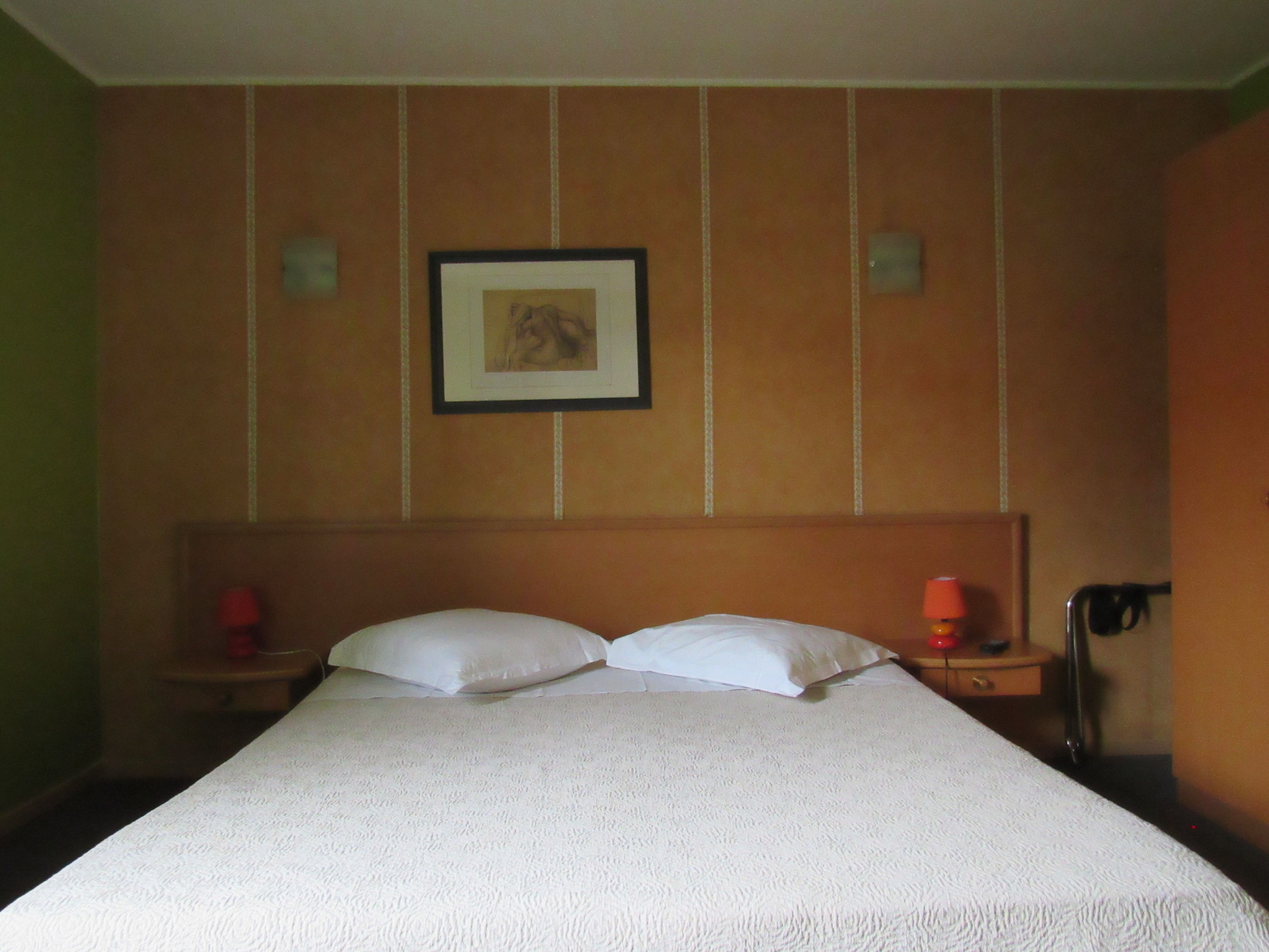 Een hotelkamer met een groot bed, twee kussens, een lijst met een afbeelding aan de muur en een nachtkastje met een lamp.