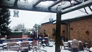 A covered outdoor seating area with wooden chairs and tables, surrounded by snow-covered plants and a brick building in the background. - Hotel In den Hoek (Tielt-Winge)