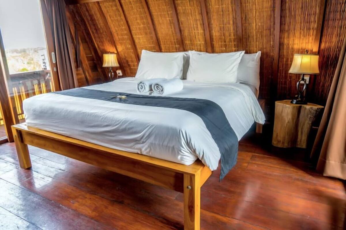 A wooden-framed bed with white and gray bedding, two bedside lamps, and a view of the outdoors.