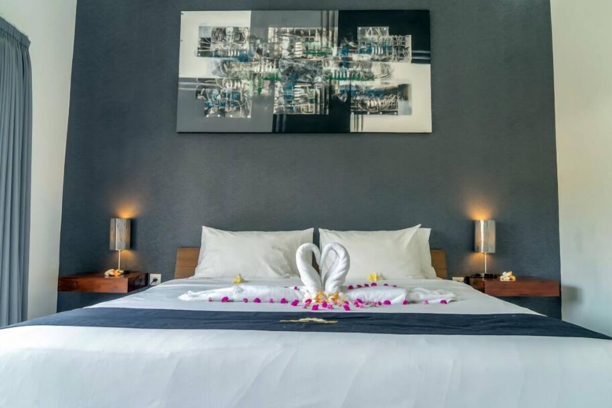 A hotel room with a large bed, two bedside tables with lamps, a wall-mounted abstract art piece, and a dark-colored headboard.