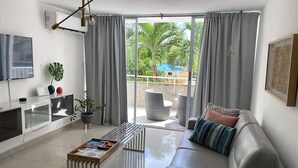 Uma sala de estar moderna com um sofá, mesa de centro e uma TV. Há uma varanda com vista para vegetação e um guarda-sol azul.