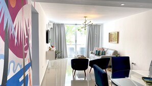 Uma sala de estar moderna com uma mesa de jantar de vidro, cadeiras azuis, um sofá e uma televisão na parede.