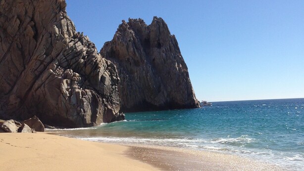 A beach with clear blue water, sandy shore, and rocky cliffs. - Villa 2206 2BR/3BA Ground Level Condo at Villa La Estancia on Medano Beach (Cabo San Lucas)