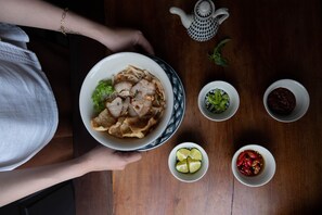A person holding a bowl of noodles with meat and vegetables, surrounded by small bowls of condiments on a wooden table. - The Mansion Hoi An by Minova (Da Nang)