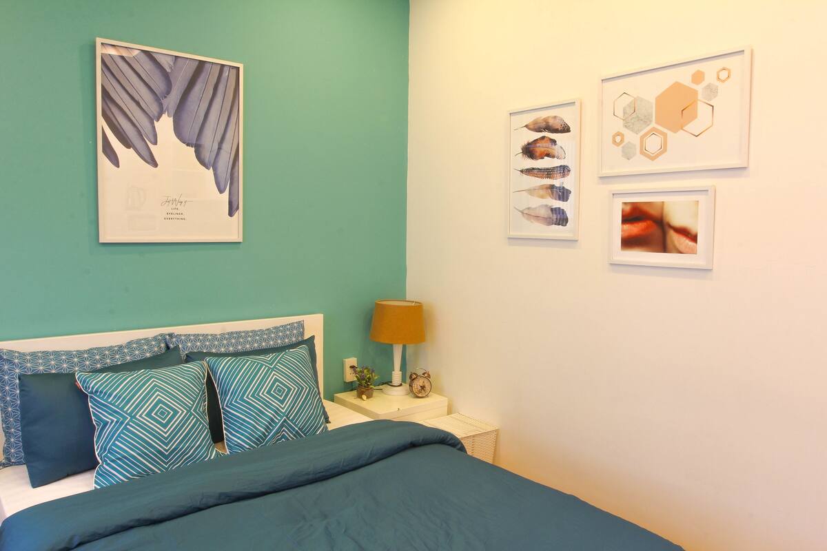 Un dormitorio con pared color turquesa, cama con almohadas estampadas, mesita de noche con lámpara y reloj, y decoración en la pared que incluye una mariposa y diseños abstractos.