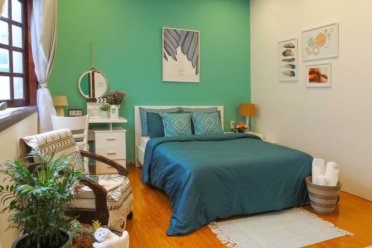 Un dormitorio con colcha color aguamarina, suelo de madera, cómoda blanca, una silla, una planta en maceta y una obra de arte en la pared.