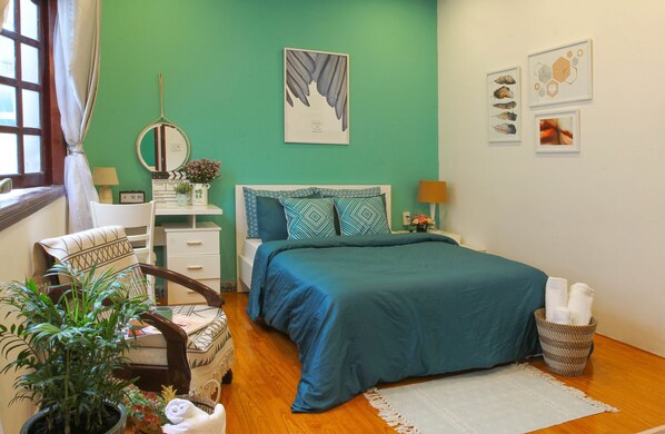 A bedroom with a teal bedspread, a wooden floor, a white dresser, a chair, a potted plant, and artwork on the wall. - Bong Gio Apartment (Ho Chi Minh City)