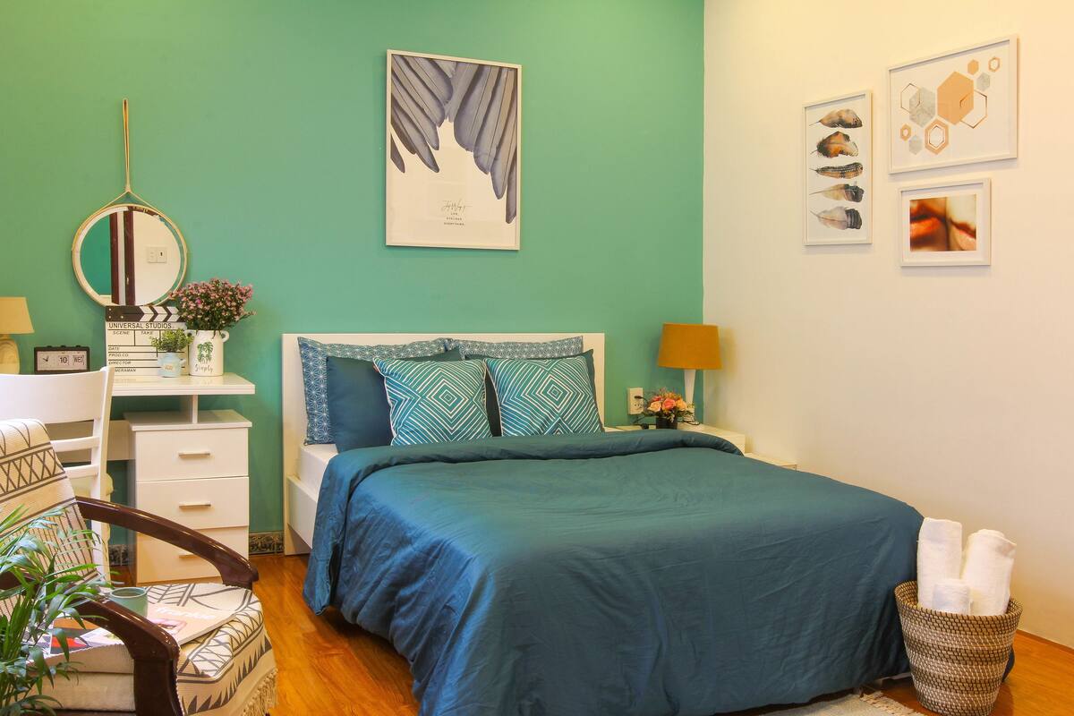 Un dormitorio ordenado con cama, mesitas de noche, una silla y una pared decorada con obras de arte.