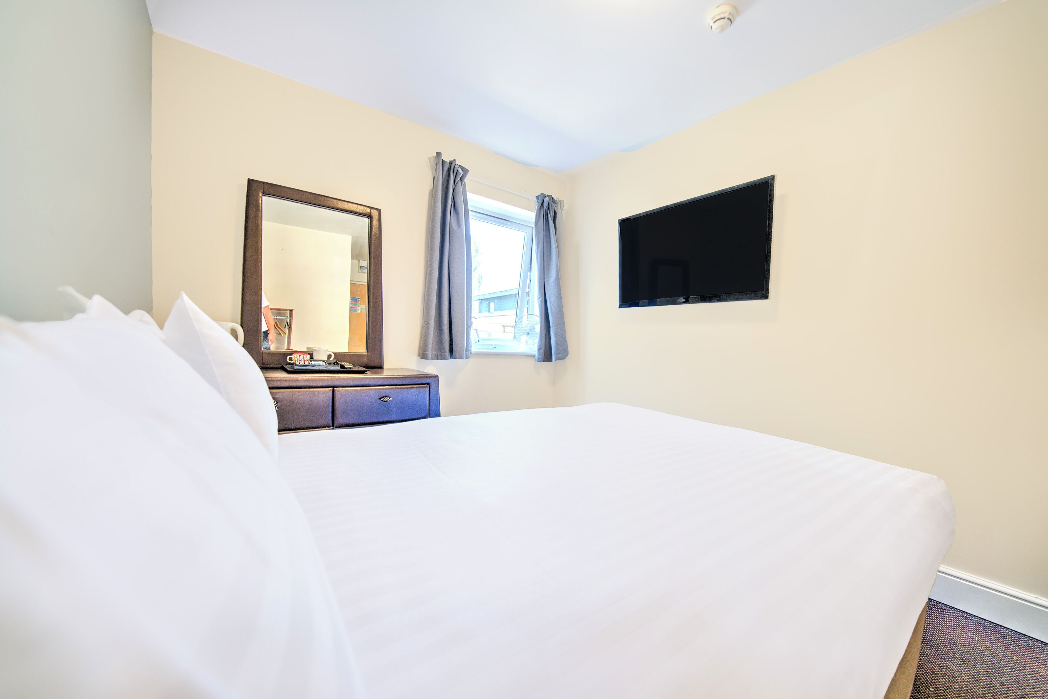 A hotel room with a large bed, a dresser, a mirror, a flat-screen TV, and a window with curtains.