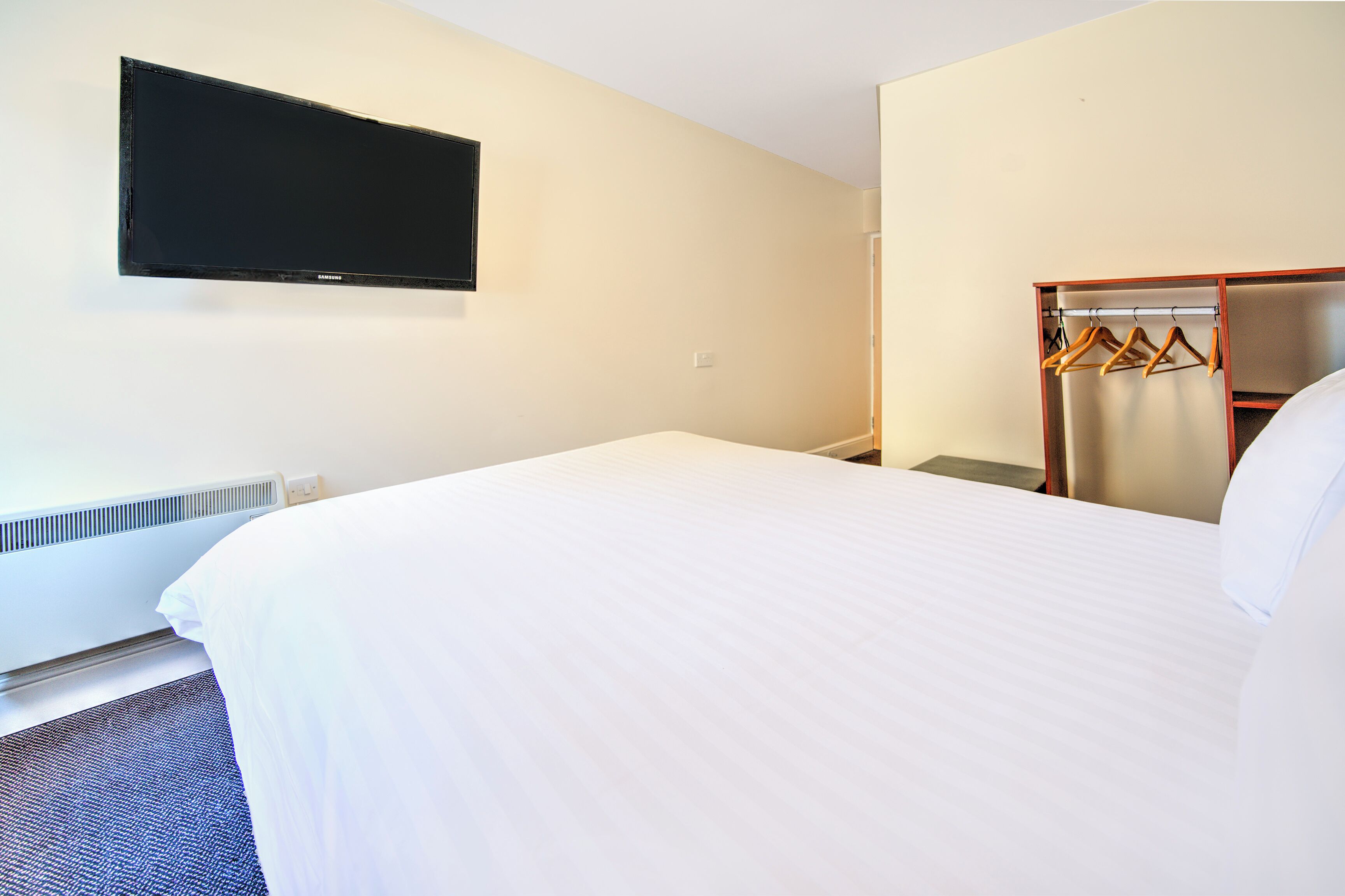 A hotel room with a large bed, a flat-screen TV mounted on the wall, a wardrobe with wooden hangers, and a radiator under the bed.