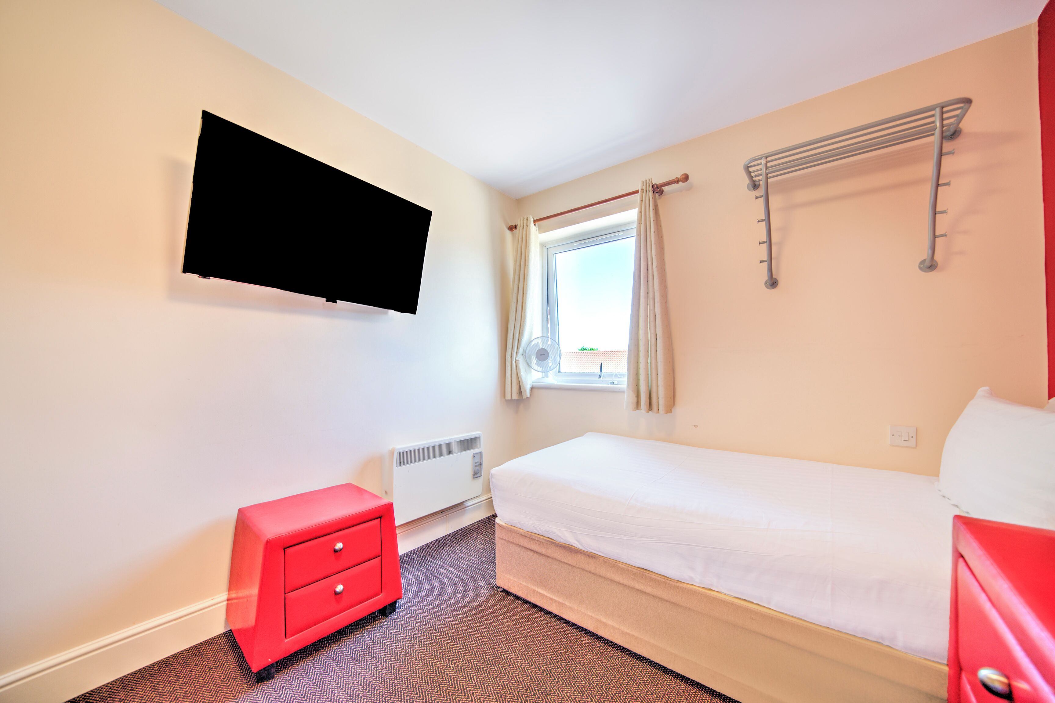Une petite chambre propre, avec un lit, une commode rouge, un téléviseur fixé au mur et une fenêtre avec des rideaux.
