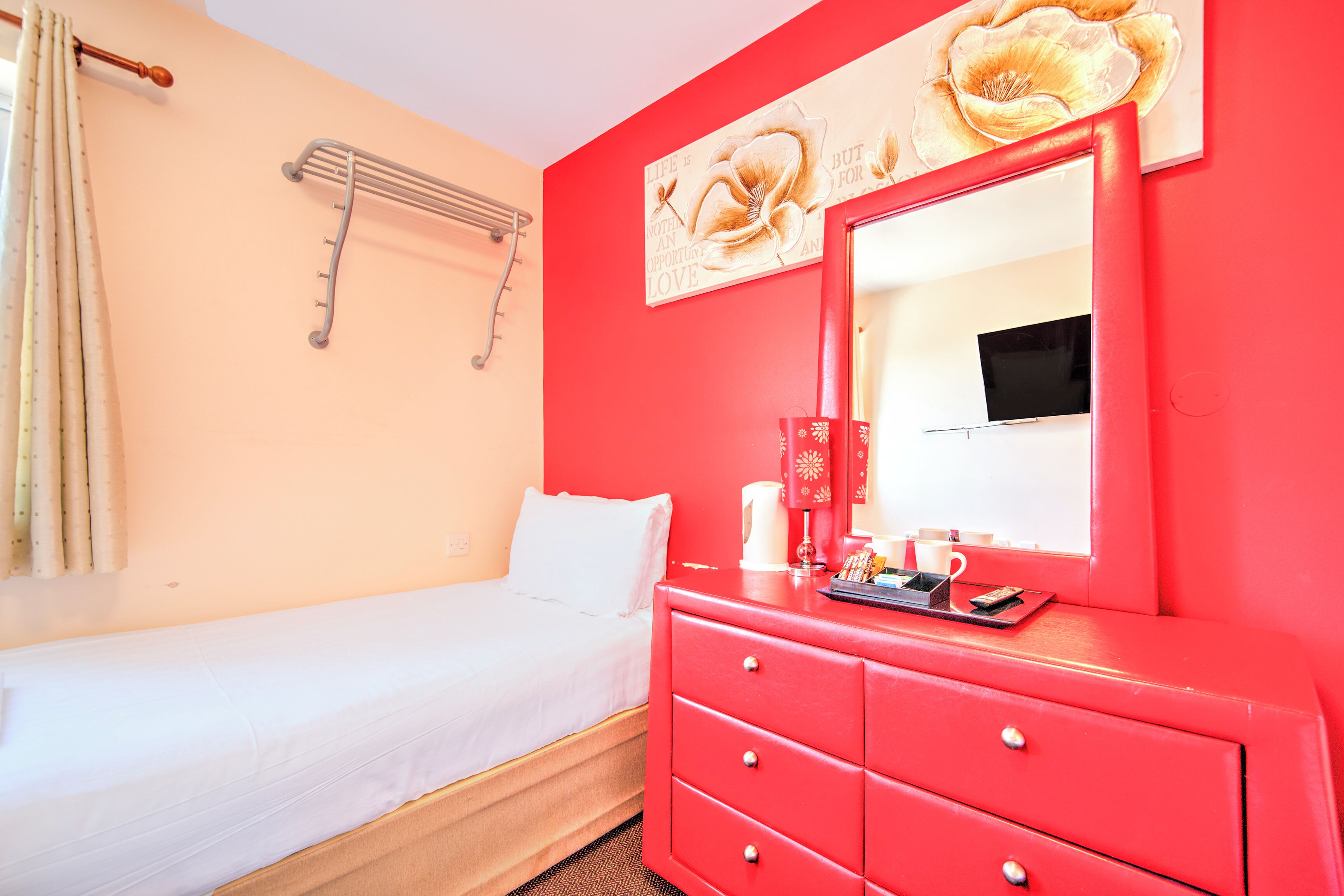 Habitación de hotel con una cama, un tocador rojo, un espejo y una pared decorada con motivos florales.