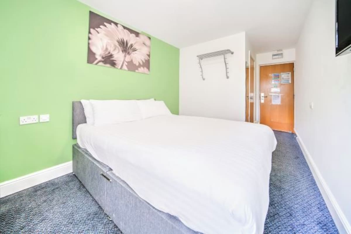A hotel room with a bed, green walls, a flower painting, a towel rack, and a door with a sign.