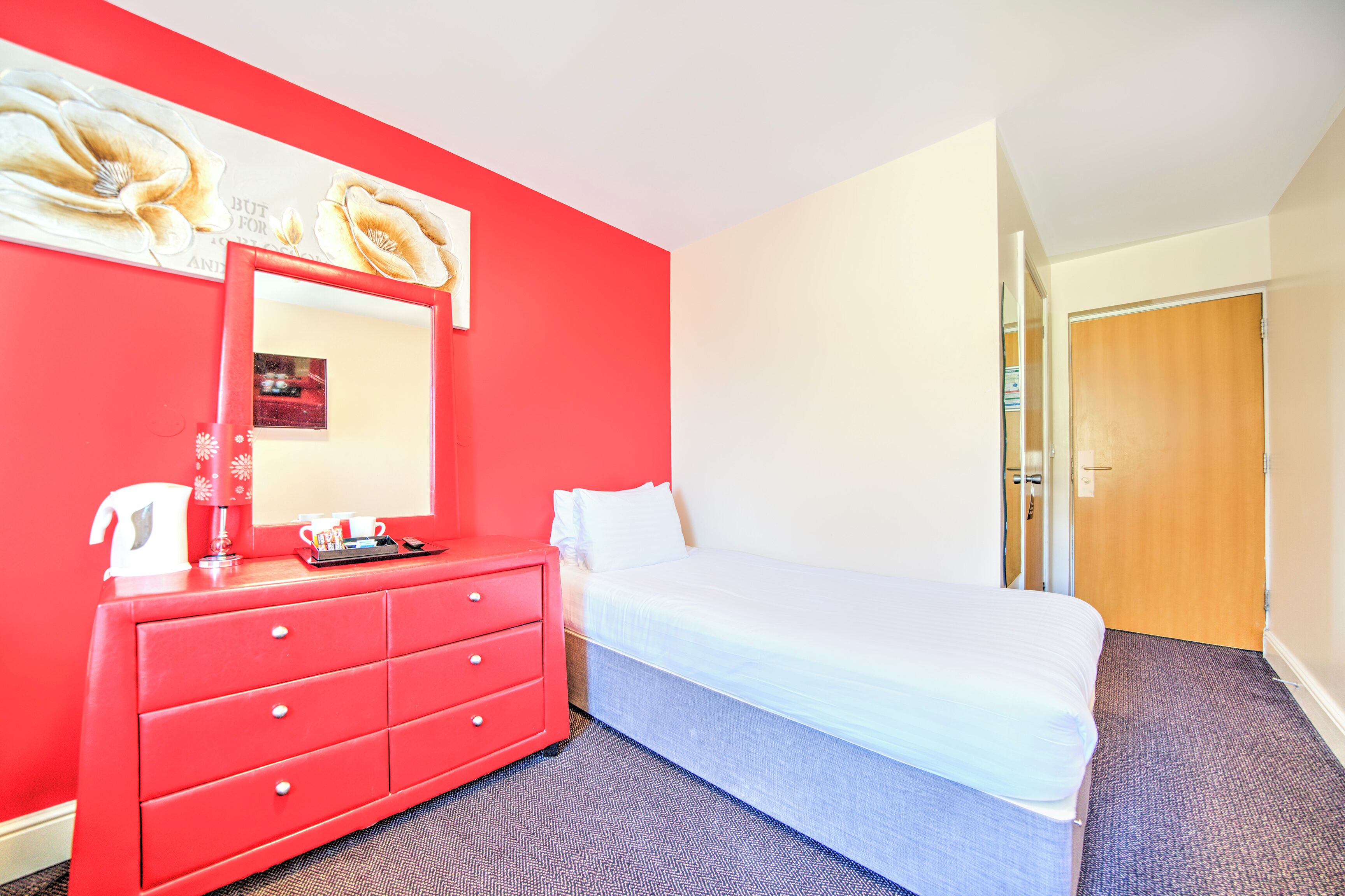 Una habitación de hotel con un aparador rojo, una cama, un espejo grande y una puerta que da a otra habitación.