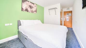 A hotel room with a bed, green walls, a flower painting, a towel rack, and a door with a sign.