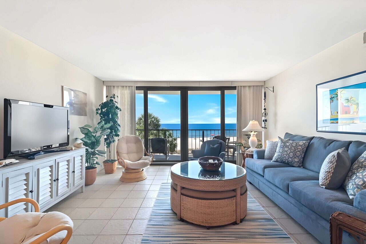 A living room with a television, sofa, chairs, a round table, and a large window with a view of the beach.
