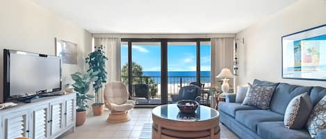 A living room with a television, sofa, chairs, a round table, and a large window with a view of the beach.