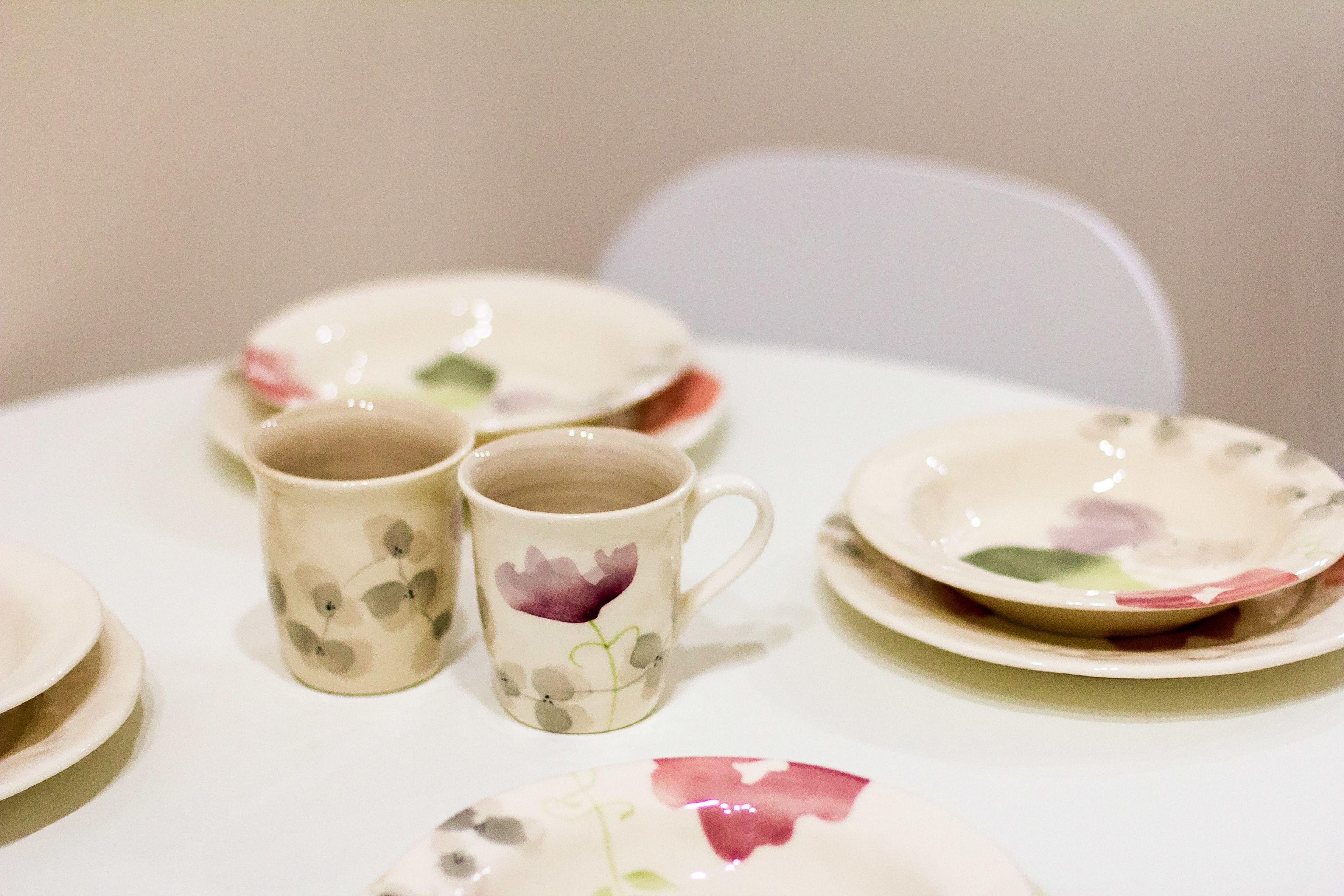 A table with a white tablecloth, a white chair, and a floral-patterned plate and mug.