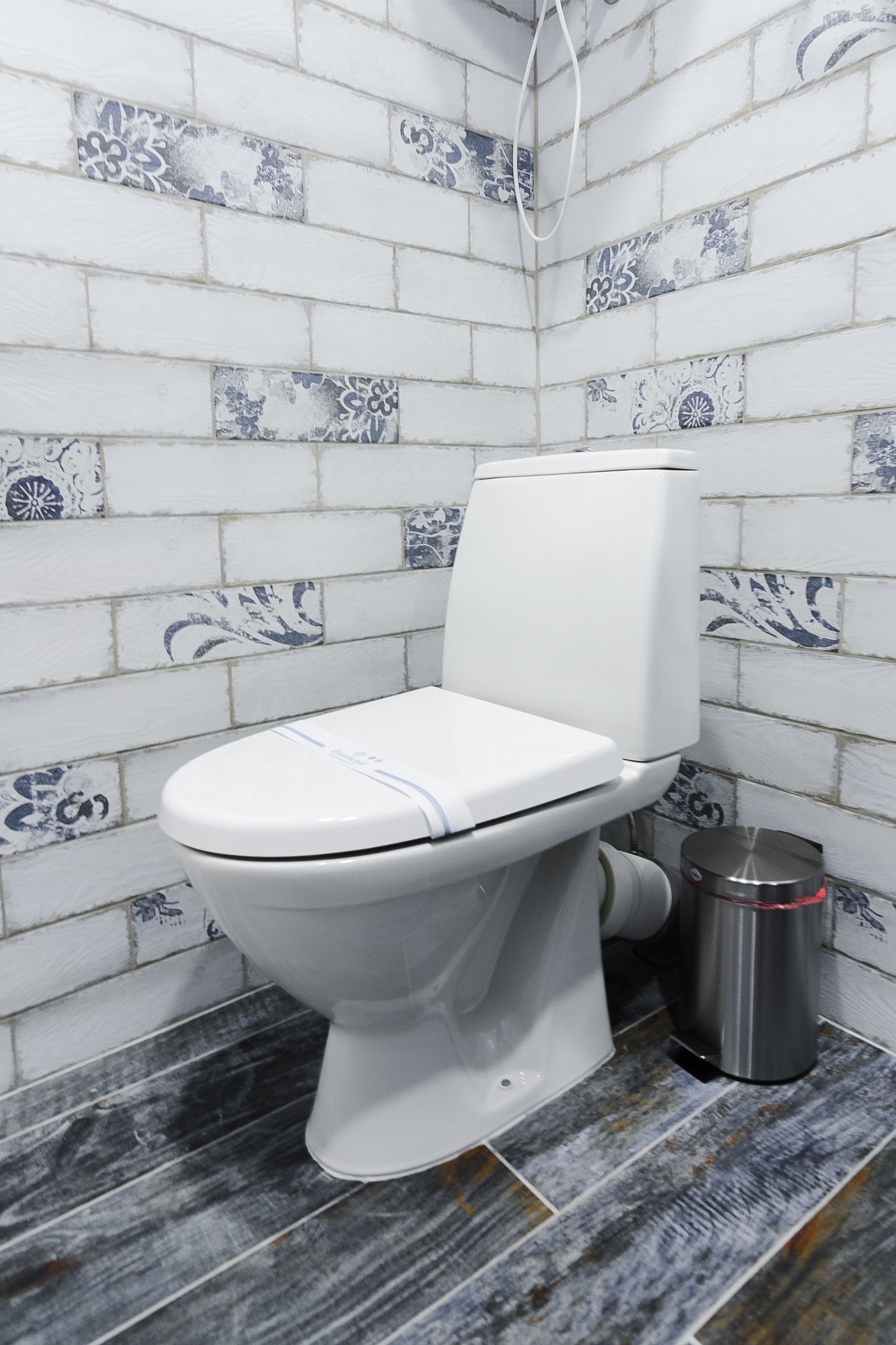 Un baño con un inodoro blanco, un cubo de basura y azulejos con diseños en las paredes.