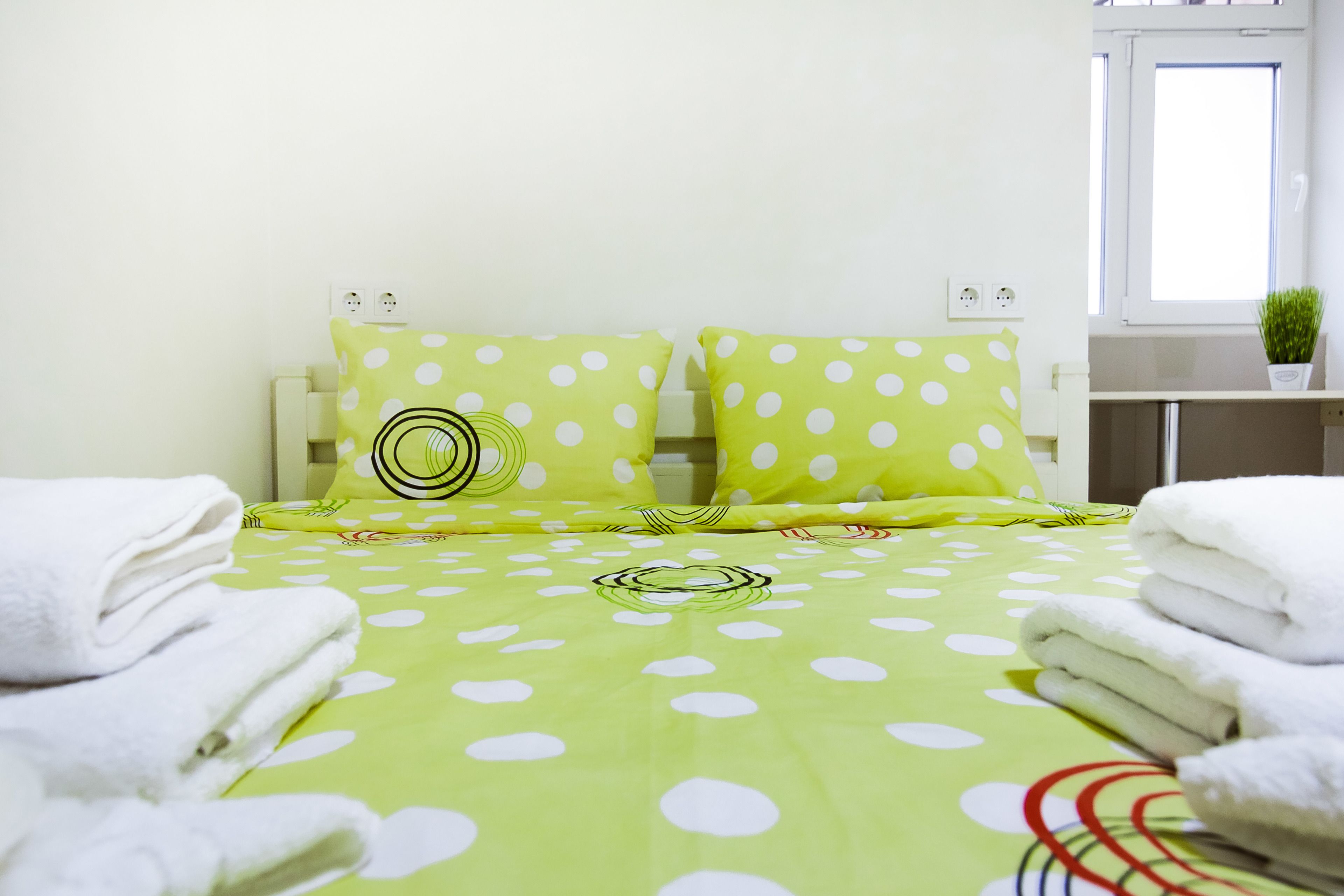 A neatly made bed with a green polka dot bedspread, two matching pillows, and folded white towels on either side.