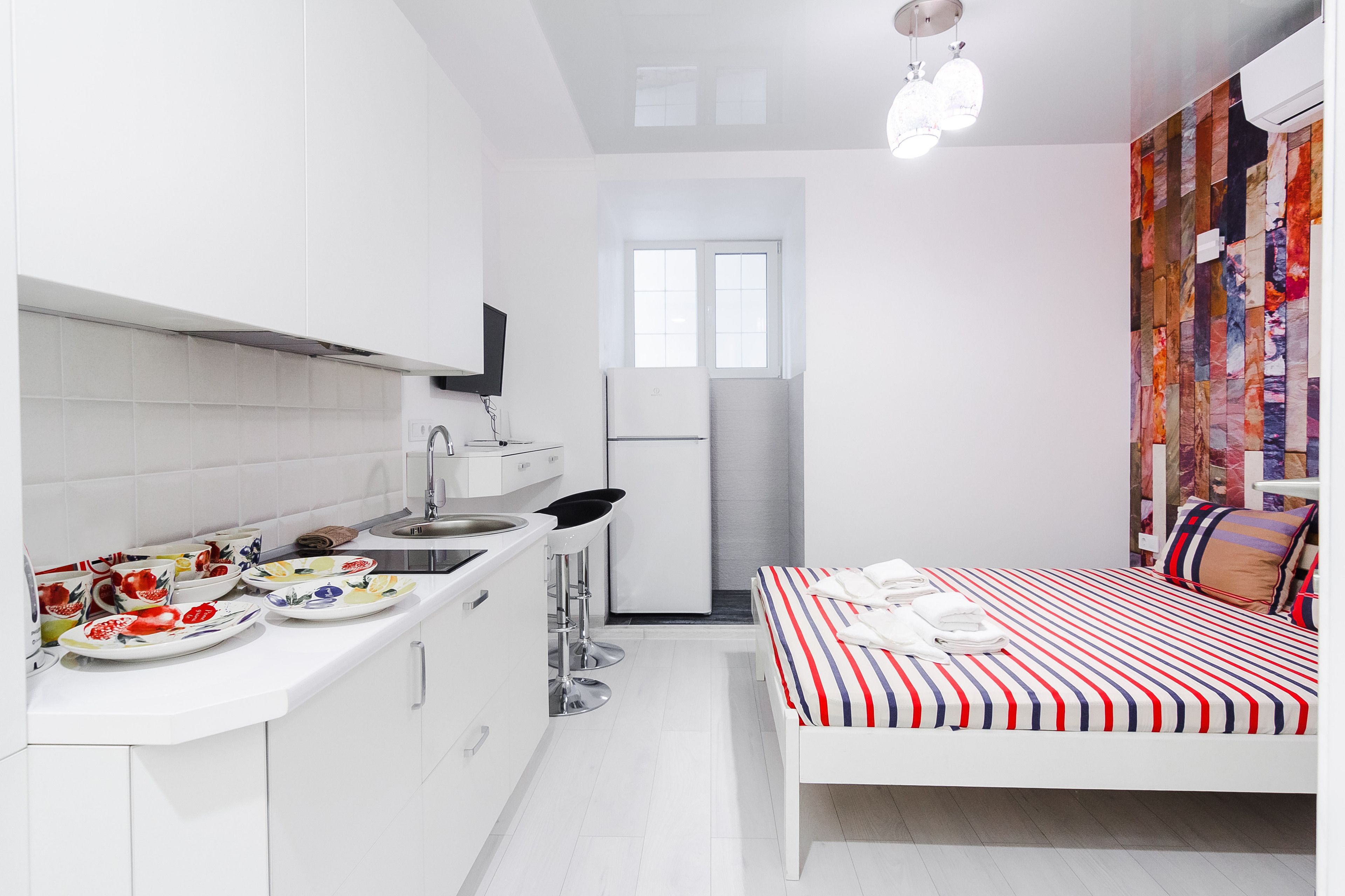 A modern kitchen with white cabinets, a sink, and a refrigerator. There is a bed with a striped bedspread and a window with a view of the city.