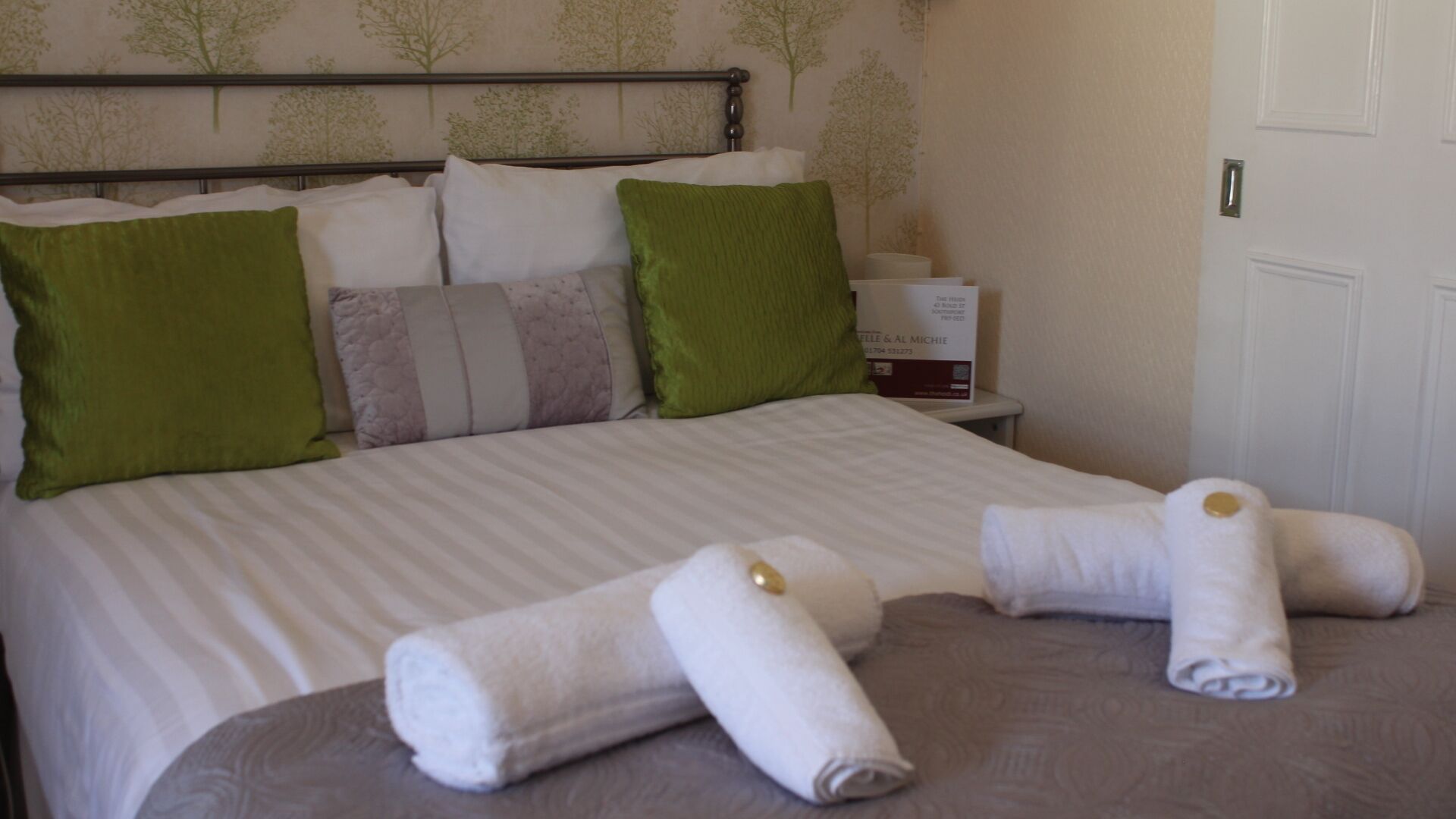 A neatly made bed with a striped bedspread, green and white pillows, and two rolled towels on the bed.