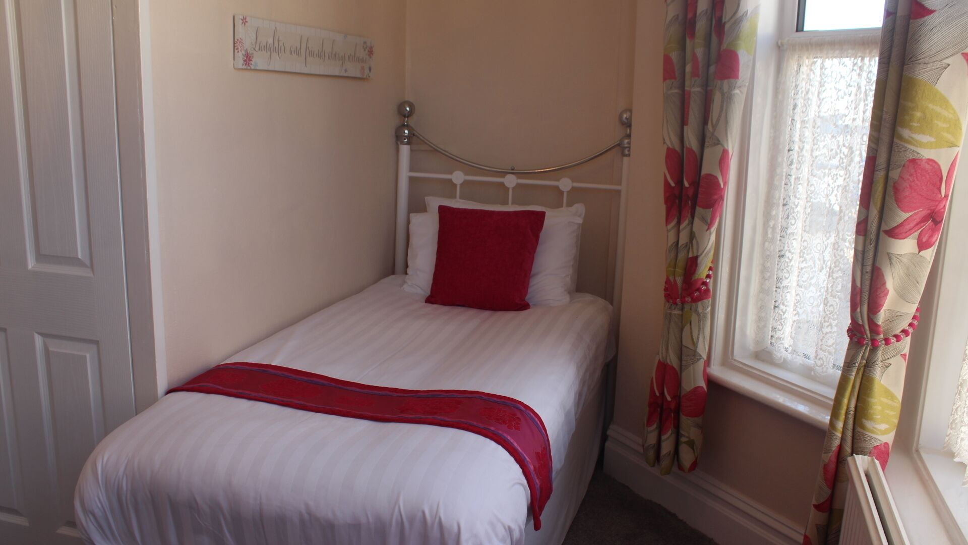 A single bed with white bedding and a red pillow, a decorative wall sign, and a window with floral curtains.