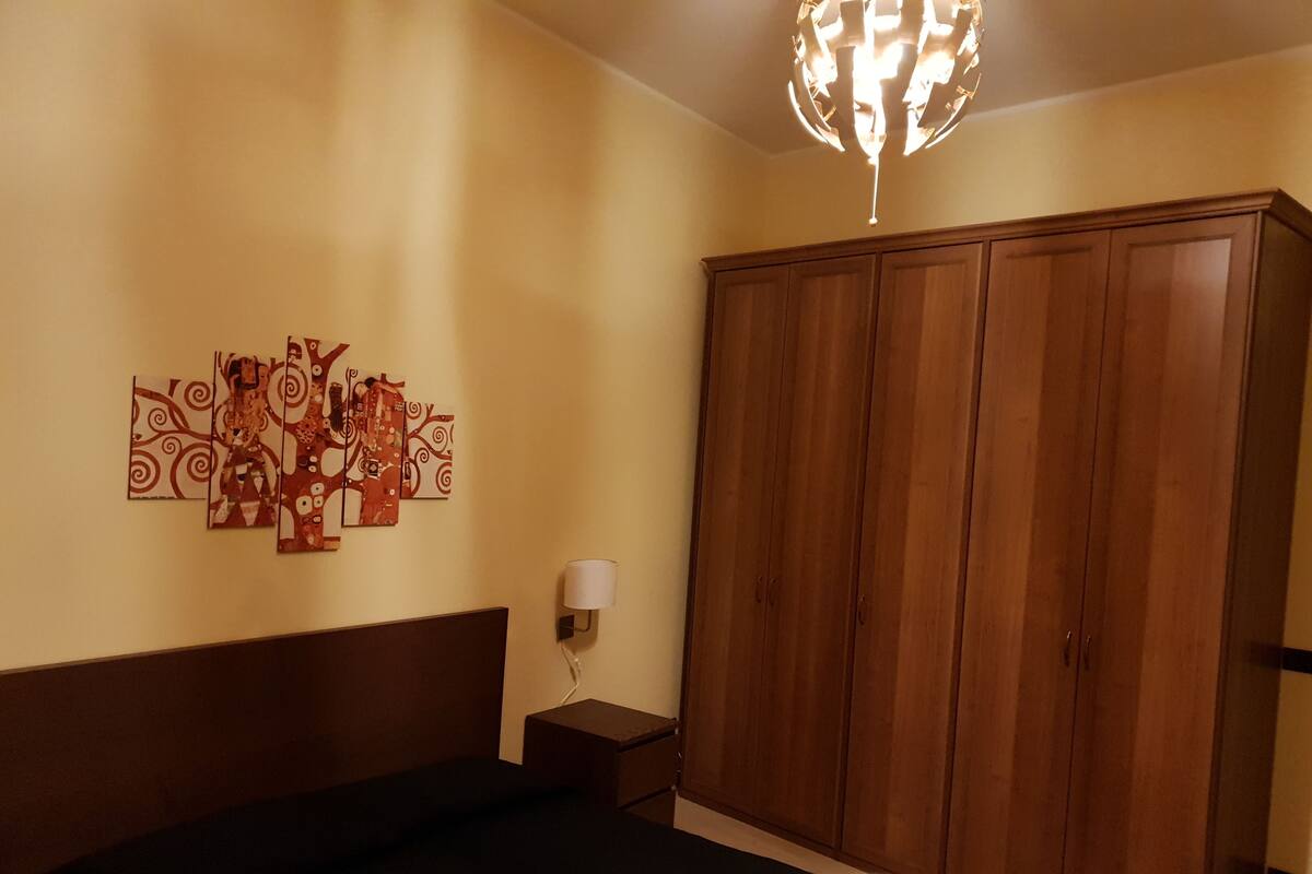 A hotel room with a bed, a wooden wardrobe, a wall-mounted lamp, and a decorative piece above the bed.
