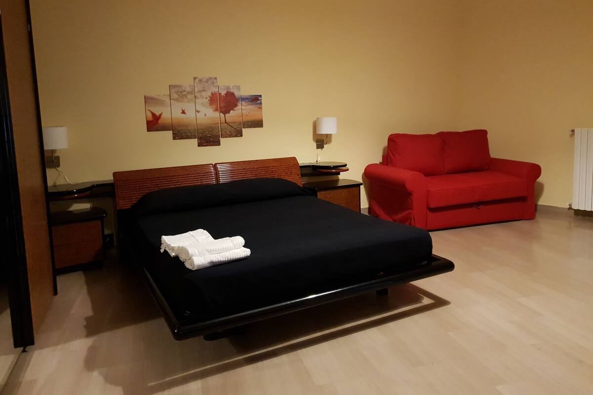 A hotel room with a bed, a red armchair, a nightstand with a lamp, and a wall with a painting.