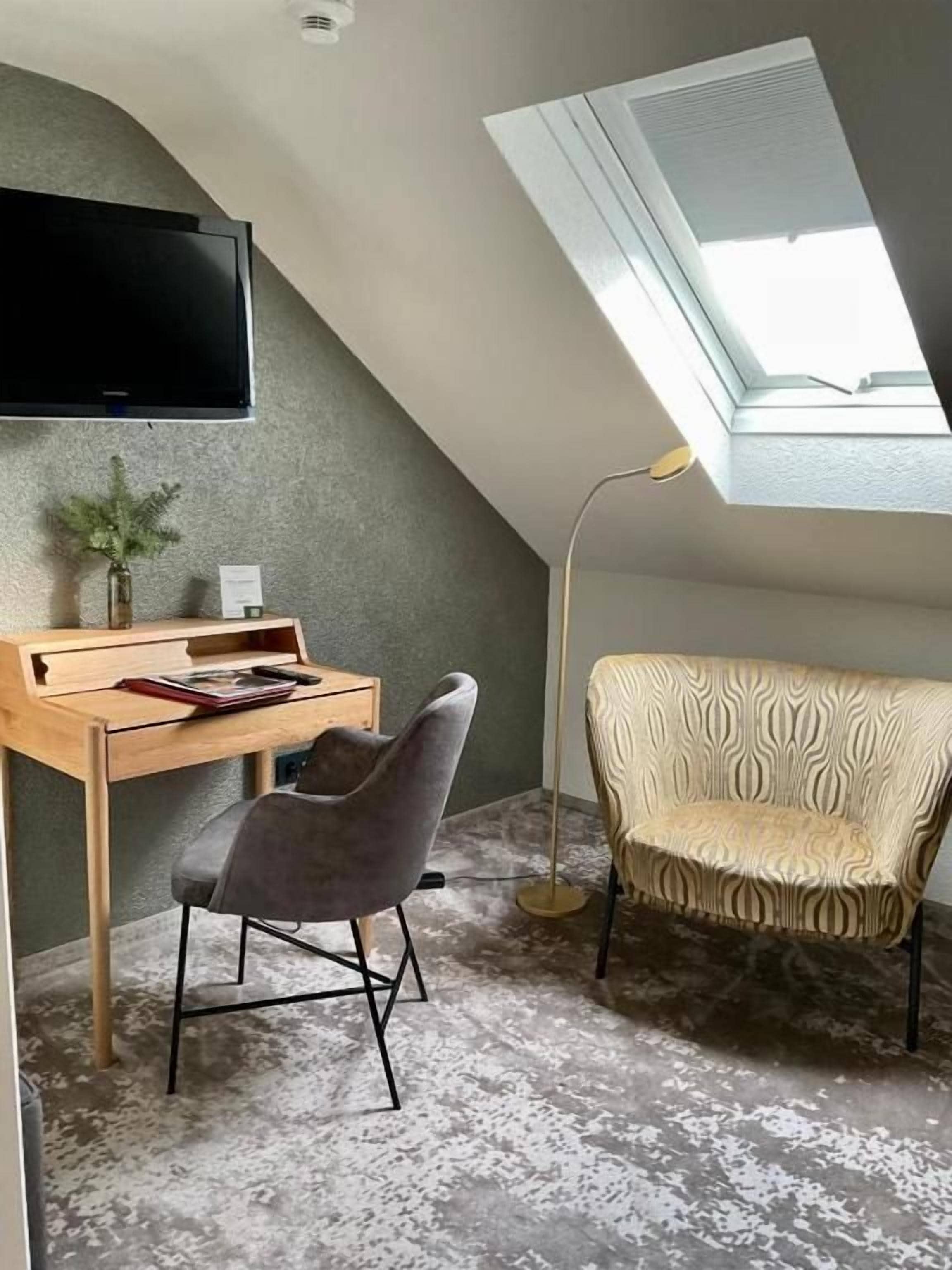 A room with a sloped ceiling, a flat-screen TV mounted on the wall, a desk with a chair, a patterned sofa, and a floor lamp.