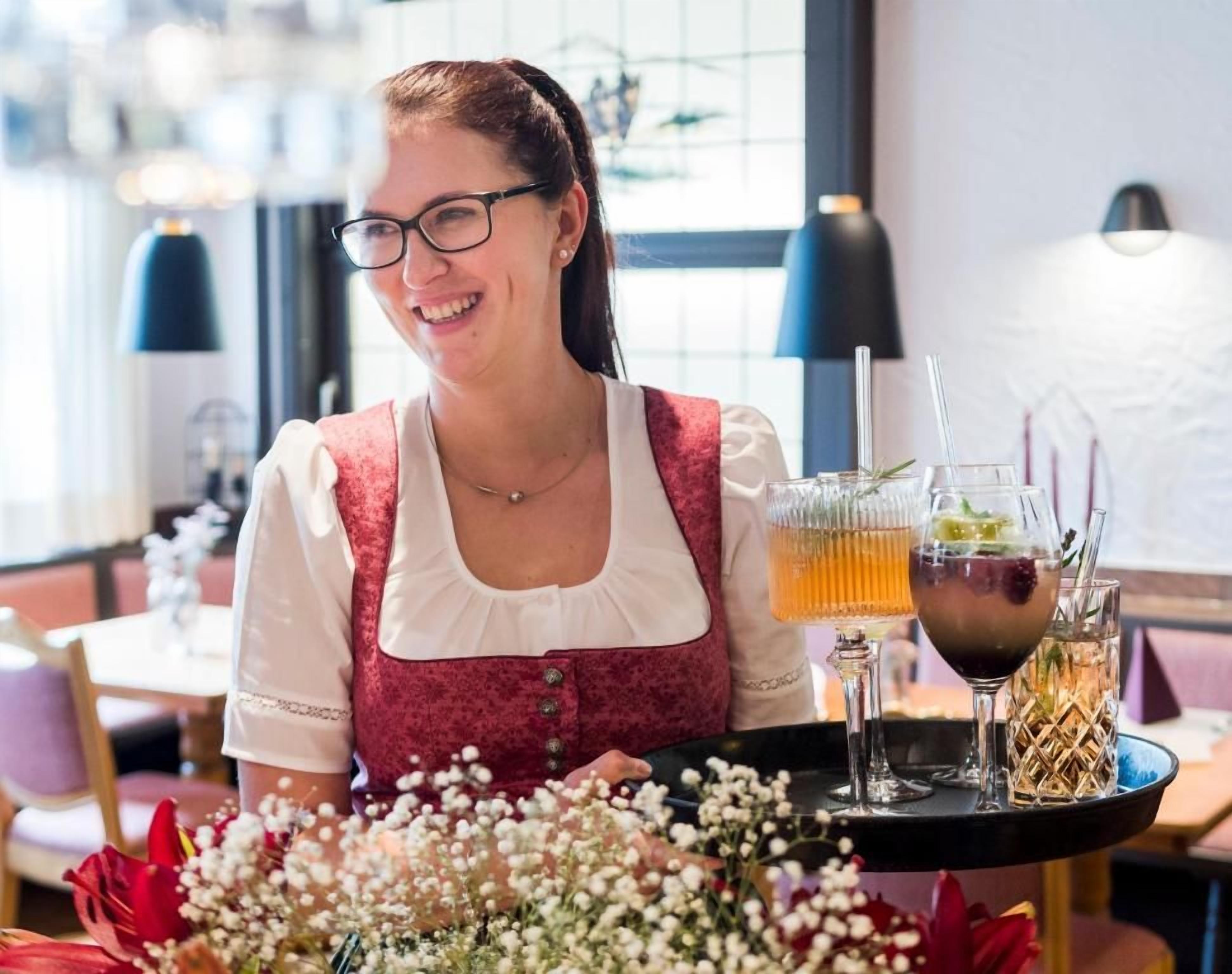 Une personne est assise à une table, devant un plateau sur lequel se trouvent une boisson et un verre. La scène se déroule dans une pièce avec une fenêtre et une lampe en arrière-plan.