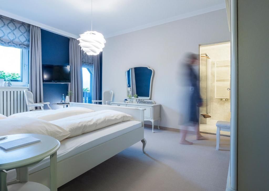 A hotel room with a bed, bedside table, and a mirror. There is a window with curtains, a chair, and a small table.