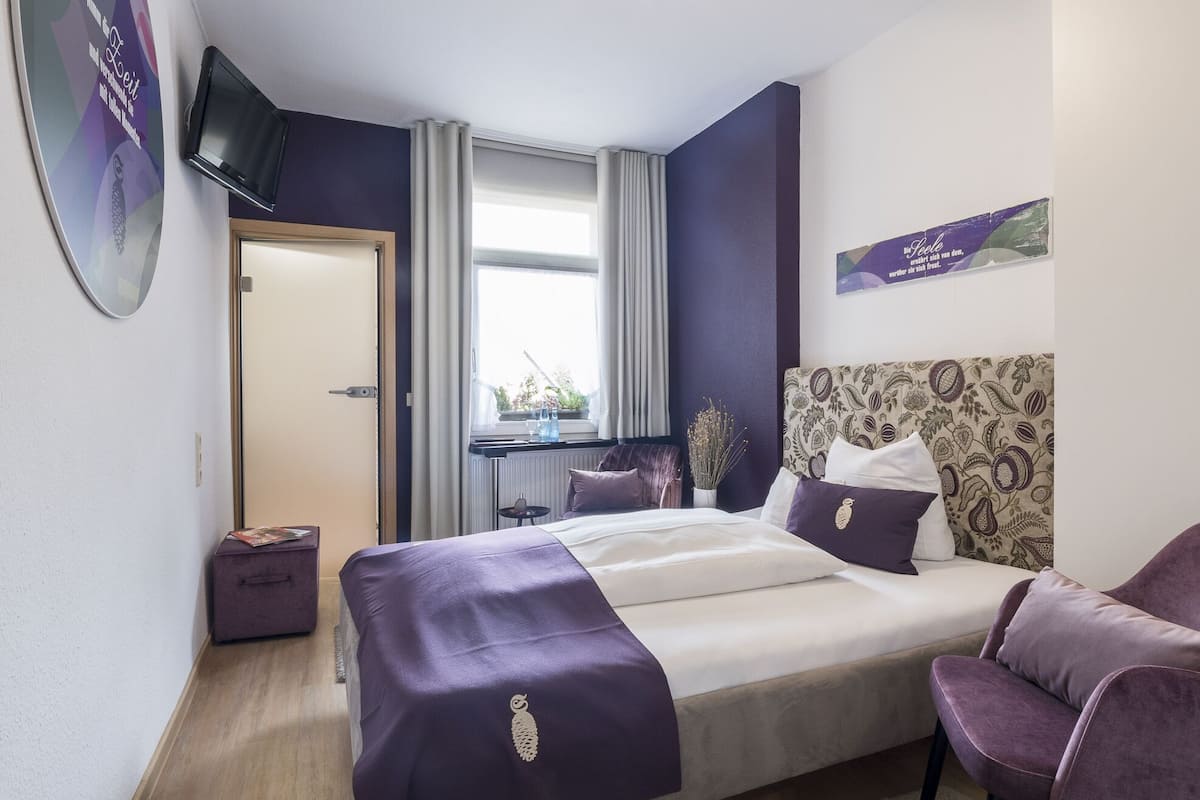 Een moderne hotelkamer met een groot bed, een aan de muur bevestigde televisie, een raam met gordijnen en een klein tafeltje met een plant.