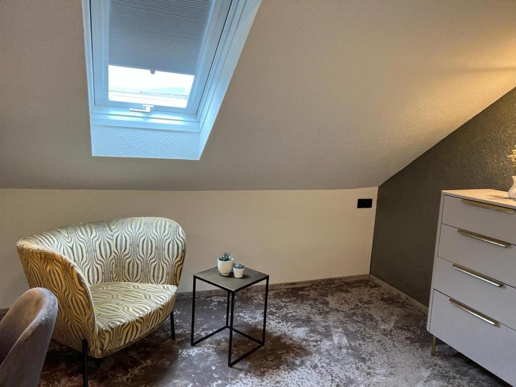 Una habitación con techo inclinado, tragaluces, un sillón estampado, una mesita con dos plantas en macetas y un aparador blanco.