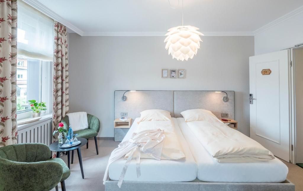 Ein Hotelzimmer mit Doppelbett, Nachttisch, Stuhl, kleinem Tisch mit Vase und Pflanze sowie einem Fenster mit Vorhängen.