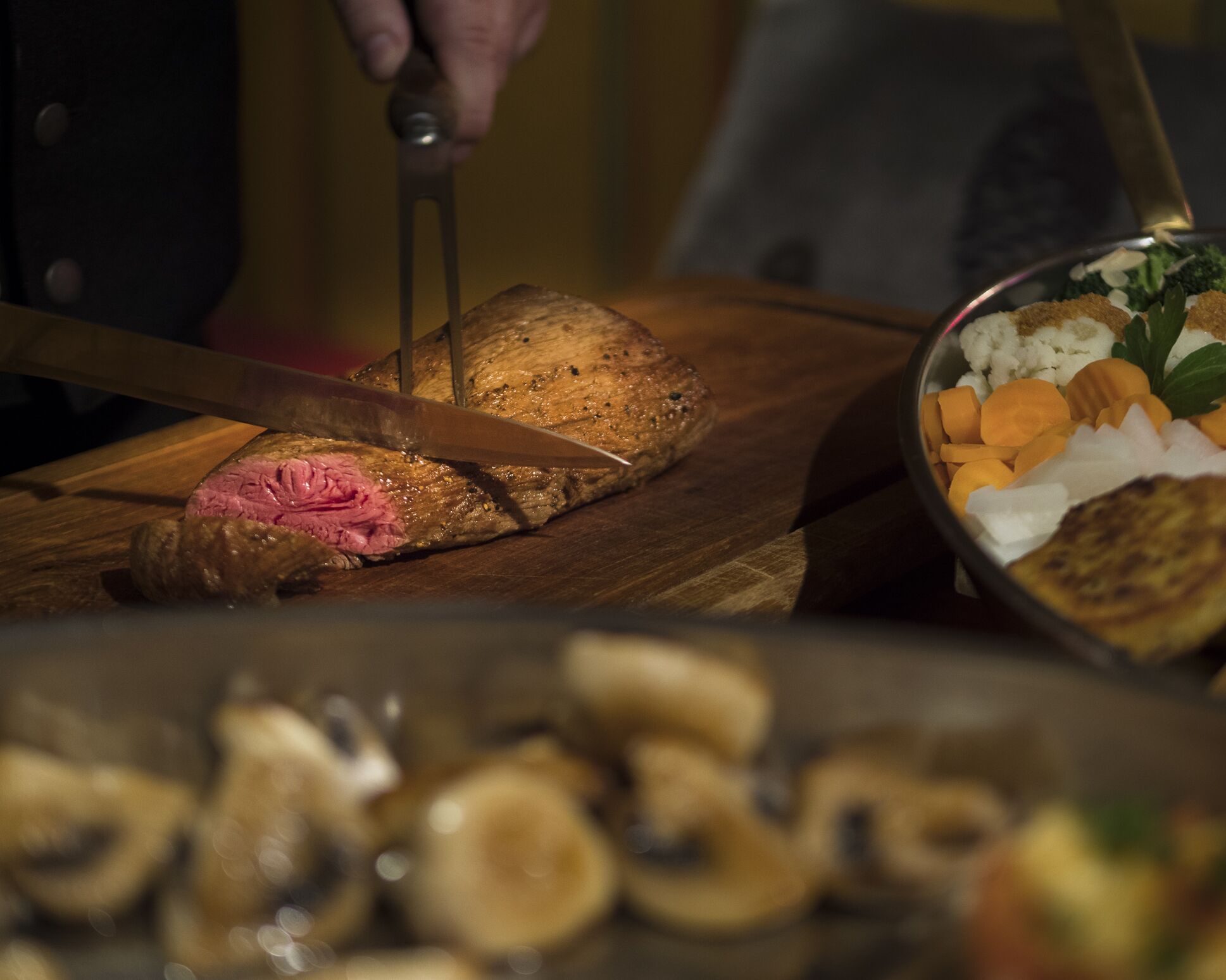 A chef slicing meat on a wooden cutting board, with a bowl of vegetables and mushrooms in the foreground.