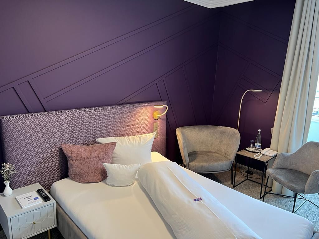 Ein Hotelzimmer mit einem großen Bett, zwei Stühlen, einem Schreibtisch mit Lampe sowie einem Nachttisch mit Telefon und kleiner Vase.