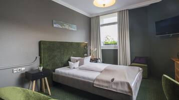 A hotel room with a bed, a green chair, a nightstand, a lamp, a window with curtains, and a television.